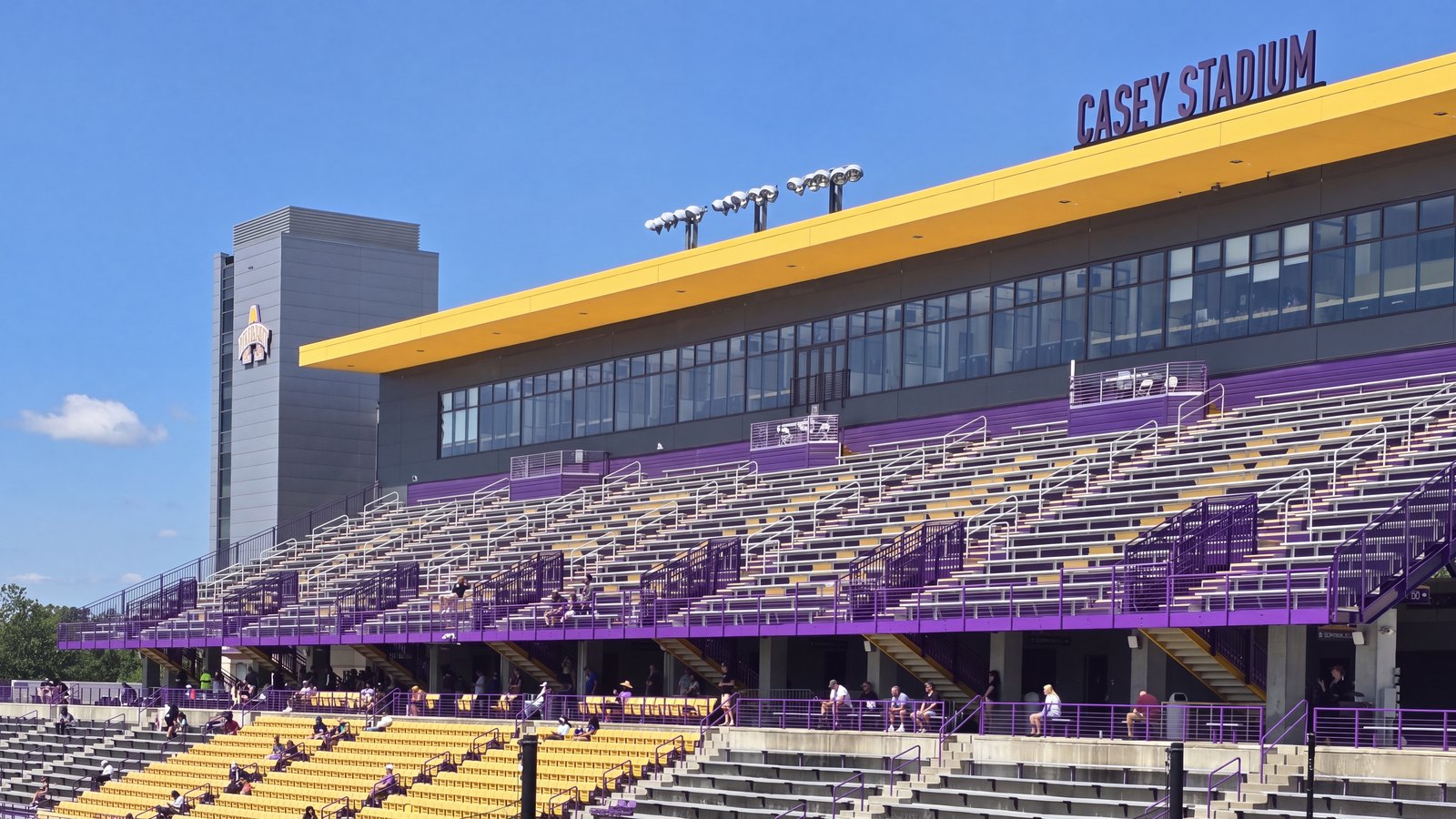 The repainted purple-and-gold grandstand at Casey Stadium, University at Albany, seen from the field under a blue sky